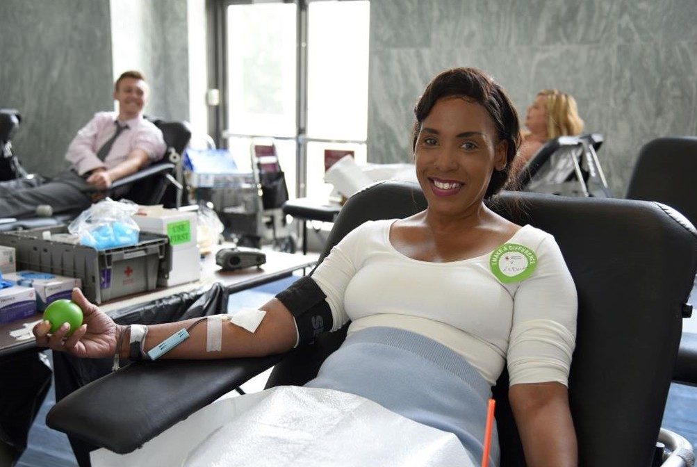 a woman is laying on a bed donating blood. she has a squeeze ball in her hand and is smiling at the camera