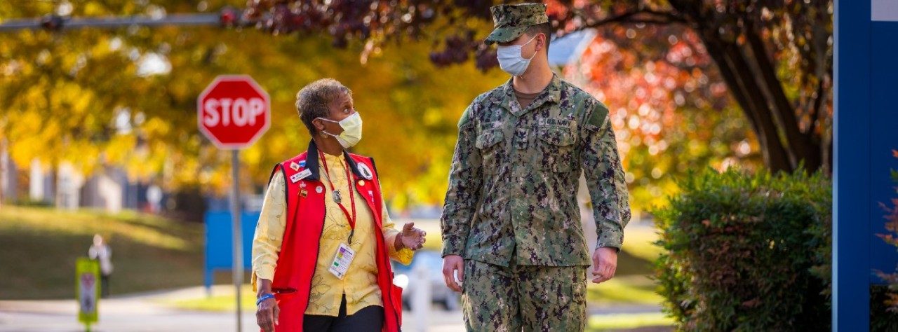 a Red Cross working in a red vest talks while walking beside a soldier wearing camoflague fatigues. they are both wearing medical face masks.