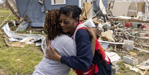 A Red Cross volunteer is hugging a women who's home was destroyed in a disaster. They are standing in the front yard of a home that has been destroyed.
