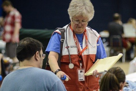 woman in red cross vest with stethoscope around neck and notepad in hand handing pill bottle to man and woman