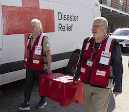 two Red Cross volunteers in red vests carry disaster supplies alongside a Red Cross truck