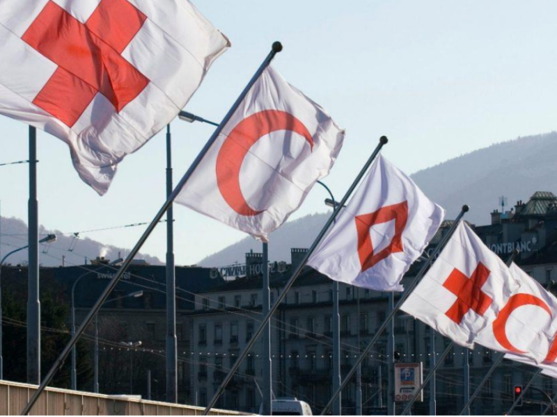 five white flags are visible in flag mounts along the edge of a building that represent the various Red Cross national societies - the cross, the crescent, and the gem are repeated emblems